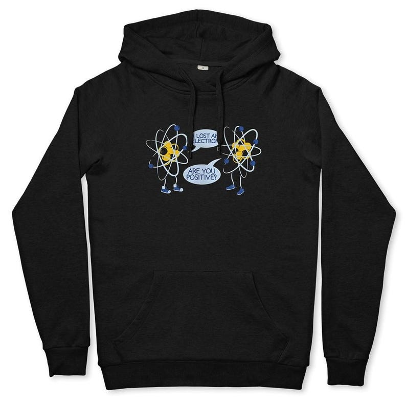 May include: Black hoodie featuring a humorous cartoon of two atoms. One atom says, "Lost an electron?" and the other replies, "Are you positive?" The design includes yellow, blue, and white accents. The hoodie has a front pocket and a hood.