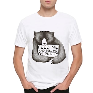 以下が含まれることがあります： 白いTシャツに、黒と白のクマのイラストが描かれています。クマは「私を餌付けして、私がきれいだと教えて」と書かれた看板を持っています。