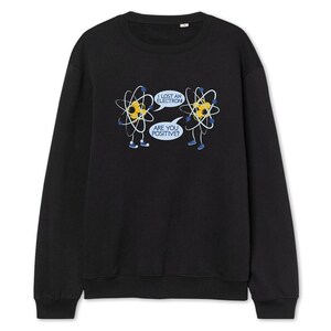May include: Black sweatshirt featuring a humorous graphic of two cartoon atoms with speech bubbles. One atom says, "I lost an electron," and the other replies, "Are you positive?" The atoms are yellow with white and black details.
