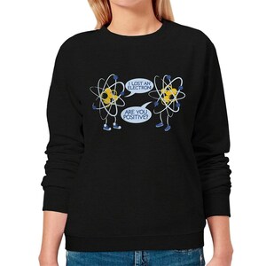 May include: Black crew neck sweatshirt featuring a humorous cartoon design of two atoms. One atom says, "I lost an electron," and the other replies, "Are you positive?" The atoms are yellow with blue accents.
