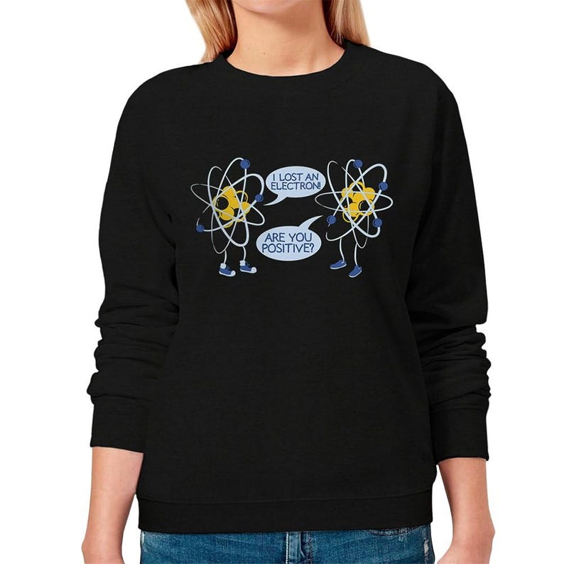May include: Black crew neck sweatshirt featuring a humorous cartoon design of two atoms. One atom says, "I lost an electron," and the other replies, "Are you positive?" The atoms are yellow with blue accents.