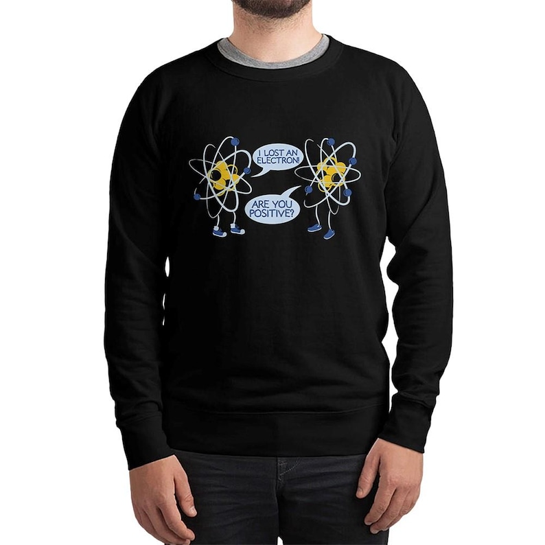 May include: Black sweatshirt featuring a humorous cartoon design of two atoms with legs and arms. One atom says, "I lost an electron," and the other replies, "Are you positive?" The design is in white and yellow.