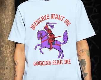 May include: A light blue t-shirt with a graphic print of a knight riding a purple horse. The text on the shirt reads "Wenches Want Me Goblins Fear Me".