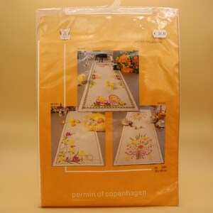 May include: A package featuring three Easter-themed table runner designs. The runners are beige with embroidered images of chicks, eggs, and floral arrangements. The package is labeled "Easter Collection" and includes the text "permin of copenhagen".