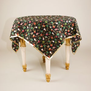 May include: A round table draped with a black Christmas tablecloth. The cloth is patterned with green Christmas trees, red and white gifts, and gold detailing. A gold border trims the cloth. The table has four white legs with gold detailing.