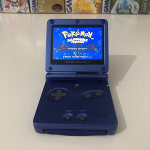 May include: A blue Game Boy Advance SP console, open to display the "Pokémon Sapphire Version" game. The screen shows the game's title and the words "Press Start". The console is on a white surface.
