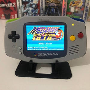 May include: A gray Nintendo Game Boy Advance handheld console displaying the "Mega Man Battle Network 3 Blue" game. The console has a black D-pad, start and select buttons, and colored action buttons. It rests on a black stand.