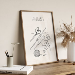 May include: A framed print titled "CROCHET ESSENTIALS" featuring line drawings of crochet tools: scissors, a crochet hook, stitch marker, needles, and a ball of yarn. The artwork is in a wooden frame and rests on a wooden surface.