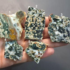 Puede incluir: Cinco piedras preciosas en bruto, sin cortar, con patrones y colores únicos. Las piedras varían en forma y tamaño, con una mezcla de tonos verdes, blancos, marrones y amarillos. Las piedras preciosas se sostienen en una mano sobre un fondo neutro. Medidas en centímetros.