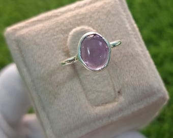 May include: A silver ring with a light purple oval-shaped gemstone. The ring is on a light pink velvet cushion.