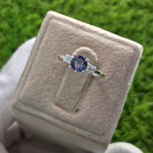 May include: A silver ring with a large, round, dark purple gemstone in the center, flanked by two smaller, clear gemstones. The ring is set in a velvet-lined jewelry box, showcasing the ring's design and sparkle.
