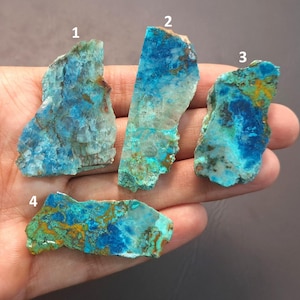 May include: Four pieces of raw chrysocolla gemstone, each with unique patterns and colours. The stones display shades of blue, turquoise, and green, with brown and gold inclusions. The rough, natural edges of the stones are visible.