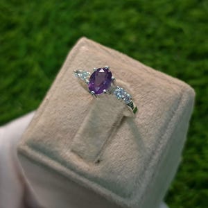 Könnte beinhalten: Ein silberner Ring mit einem violetten ovalen Edelstein und kleineren klaren Edelsteinen. Der Ring wird auf einem hellgrauen Samtringetui präsentiert.