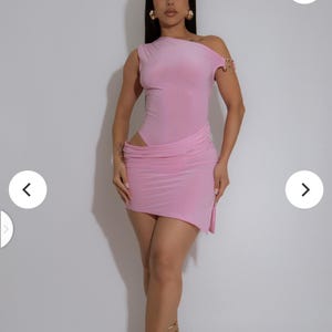May include: A light pink, one-shoulder dress with a draped skirt. The dress features a cutout at the waist and is accessorized with gold-colored arm cuffs and strappy heels. The dress is form-fitting and made of a soft fabric.