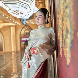 May include: A woman wearing a traditional gold-colored dress with long sleeves and a mandarin collar. The dress has a red and gold accent at the waist and a matching red tassel. The background features a decorative wall and a chandelier.