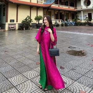 May include: A woman wearing a pink and green dress with a black purse stands in front of a traditional Asian building.