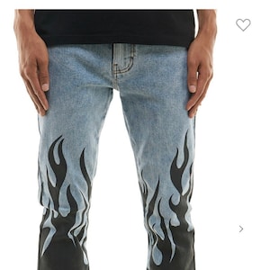 May include: Light blue denim jeans with black flame designs on the legs. The jeans have a bootcut style and are worn by a person wearing white sneakers.