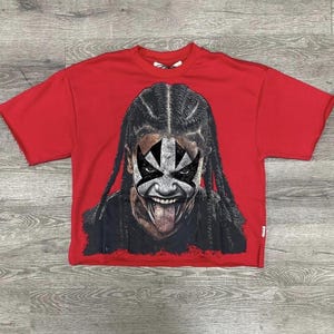 May include: Red cropped t-shirt featuring a graphic of a person with black braids and face paint. The face paint is black and white, with the tongue sticking out. The shirt has a raw hem and short sleeves.