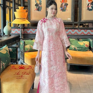 May include: A woman wearing a pink and white patterned long-sleeved dress with a high collar and a pink sash. She is standing in front of a wall with traditional Asian artwork and holding a small white purse. The text "Ao Dai -  Dù Dáng Mẹ Việt" is visible at the bottom of the image.