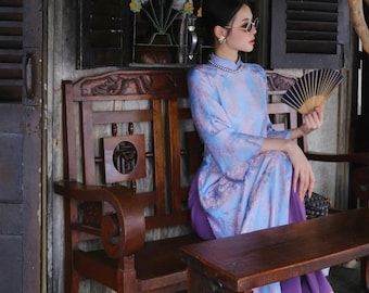 May include: A woman wearing a light blue and purple patterned dress sits on a wooden bench with a hand fan. She is wearing sunglasses and a pearl necklace.