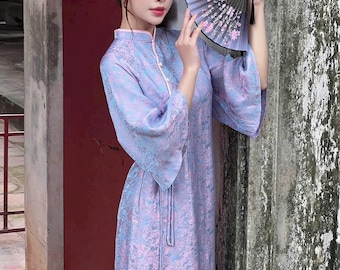 May include: A woman wearing a light blue and pink patterned dress with a high collar and long sleeves. She is holding a black and white hand fan with pink flowers.