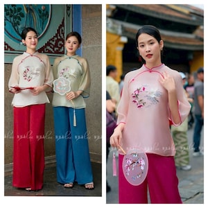 May include: Two women wearing traditional Vietnamese clothing, known as Áo dài, with embroidered designs. The women are holding fans and standing in front of a building. The Áo dài are in light pink and beige colors.