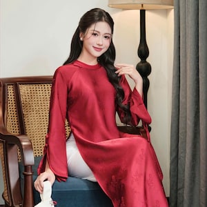 May include: A long, flowing red dress with long sleeves and a high side slit. The dress has a subtle floral pattern and is paired with white pants. The sleeves are tied at the wrists. The image also shows a white clutch and a wooden chair.