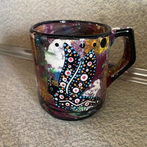 May include: A ceramic mug with a colorful, abstract design. The mug features a black handle and rim, and a butterfly illustration with blue, red, and white dots. The background has a mix of colors including purple, yellow, and green.