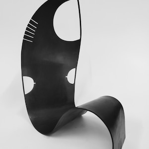 May include: A black, abstract sculpture with a curved, flowing design. The sculpture features white cut-out shapes and lines, including a large oval and two smaller, eye-like shapes. The piece stands on a white surface.