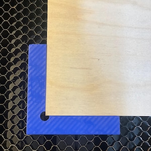 Puede incluir: Un ángulo recto de plástico azul con un agujero en la esquina, colocado sobre una superficie de rejilla negra junto a una pieza de madera marrón claro.