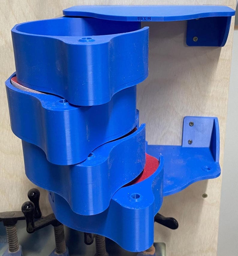 May include: A blue, 3D-printed, multi-tiered storage unit with a unique, curved design. The unit is mounted on a wooden surface and includes a shelf. The text "USH" is visible on the top. The unit appears to be designed for organizing small items.