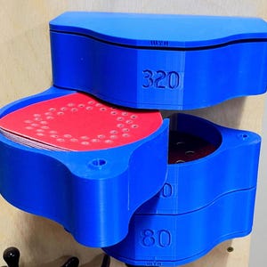 May include: A blue, 3D-printed storage container with three compartments, each labeled with numbers. The top compartment is open, revealing red circular sanding discs. The container is mounted on a wooden surface.