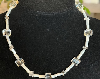 May include: A white and clear crystal beaded necklace with square-shaped beads. The necklace is on a black mannequin.
