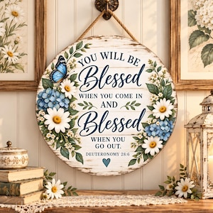 Puede incluir: Un letrero de pared redondo de madera con acabado blanco desgastado, con detalles florales azules y blancos, una mariposa azul y el texto "YOU WILL BE Blessed WHEN YOU COME IN AND Blessed WHEN YOU GO OUT. DEUTERONOMY 28:6" con un pequeño corazón azul.