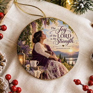 Puede incluir: Un adorno navideño redondo de cerámica con una ilustración impresa de una persona sentada al aire libre al atardecer con una taza y un libro. El texto dice The Joy of the LORD is My Strength NEHEMIAH 8:10. Incluye un cordón dorado para colgar.