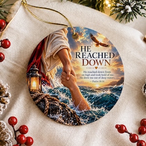 May include: A round ceramic ornament featuring a printed image of a hand reaching down to pull another from turbulent ocean waters. The text reads HE REACHED DOWN and includes the Bible verse Psalm 18:16. It has a gold hanging string.