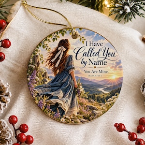 Può includere: Un ornamento rotondo in ceramica con un'illustrazione stampata di una persona dai capelli lunghi con un abito blu in un paesaggio montano. Il testo recita I Have Called You by Name You Are Mine. ISAIAH 43:1. Include un cordino dorato.