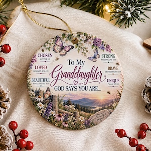 May include: A round ceramic ornament with a gold string featuring a landscape illustration of mountains at sunrise, butterflies, and wildflowers. The text reads: To My Granddaughter GOD SAYS YOU ARE... CHOSEN, LOVED, BEAUTIFUL, STRONG, BRAVE, UNIQUE.