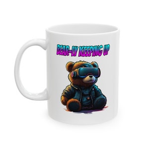 Könnte beinhalten: Weiße Keramiktasse mit einem Comic-Bären, der eine VR-Brille und eine Lederjacke trägt. Der Text "BEAR-LY KEEPING UP" ist auf die Tasse gedruckt.