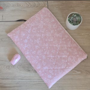 Könnte beinhalten: Eine rosa gesteppte Stofftasche mit weißem Blumenmuster, ein kleiner rosa Gegenstand und eine kleine Topf-Sukkulente auf einer Holzoberfläche. Die Tasche hat einen Reißverschluss.