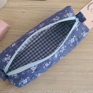 Puede incluir: Un estuche de tela acolchada en color azul polvoriento con un estampado floral blanco. El estuche cuenta con una cremallera azul claro y un forro interior con un patr&oacute;n de cuadros vichy en azul y blanco. Incluye una etiqueta con el texto ARMESETSY.