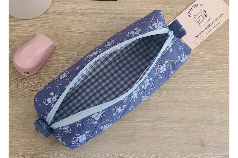 Puede incluir: Un estuche de tela acolchada en color azul polvoriento con un estampado floral blanco. El estuche cuenta con una cremallera azul claro y un forro interior con un patr&oacute;n de cuadros vichy en azul y blanco. Incluye una etiqueta con el texto ARMESETSY.