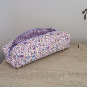 A floral patterned pencil case with a purple zipper is placed on a wooden surface next to a potted plant.