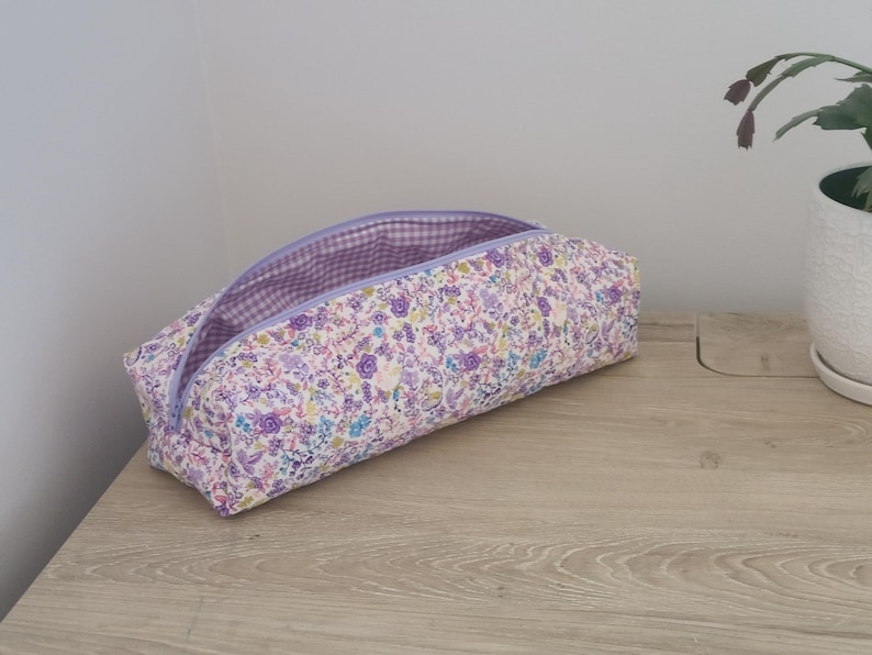 A floral patterned pencil case with a purple zipper is placed on a wooden surface next to a potted plant.
