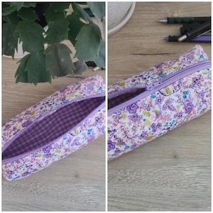Puede incluir: Un estuche cil&iacute;ndrico con un estampado floral de flores moradas, rosas y amarillas sobre un fondo blanco. El estuche tiene una cremallera morada y un forro interior de tela con cuadros vichy en tonos morados y blancos.