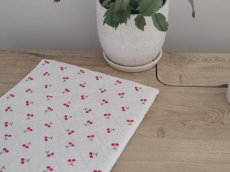 a white ceramic pot with a plant, a white cloth with red cherries, and a wooden surface.