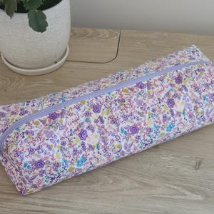 A floral patterned pencil case is placed on a wooden surface next to a white pot with a plant.