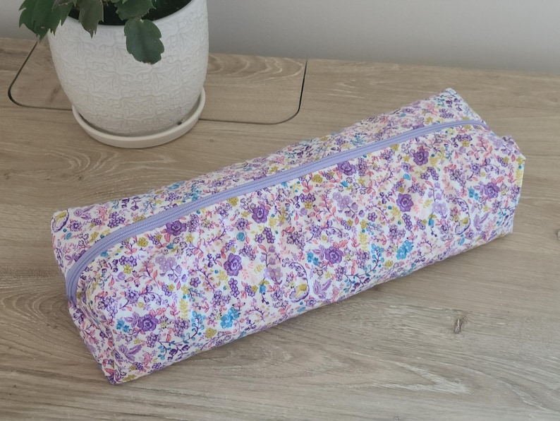 A floral patterned pencil case is placed on a wooden surface next to a white pot with a plant.