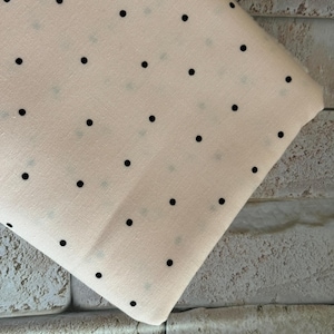 May include: A folded piece of light cream-colored fabric featuring a repeating pattern of small, solid black polka dots arranged in a grid-like formation across the surface of the textile material.