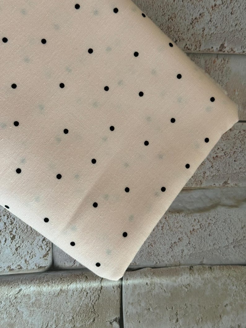 May include: A folded piece of light cream-colored fabric featuring a repeating pattern of small, solid black polka dots arranged in a grid-like formation across the surface of the textile material.